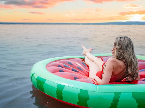 A woman relaxes on a watermelon-shaped inflatable raft in calm water at sunset, legs up, enjoying the serene lake scene.