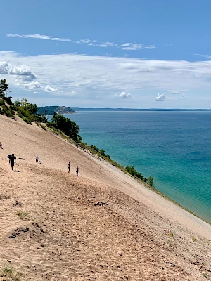 A sandy dune slopes down to a turquoise sea under a blue sky, with a few people walking along the shore and green vegetation on the ridge.
