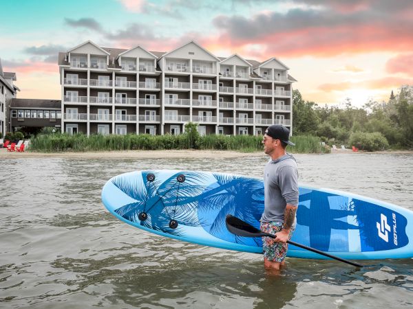 A man stands in shallow water holding a blue surfboard near a riverside hotel complex at sunset.