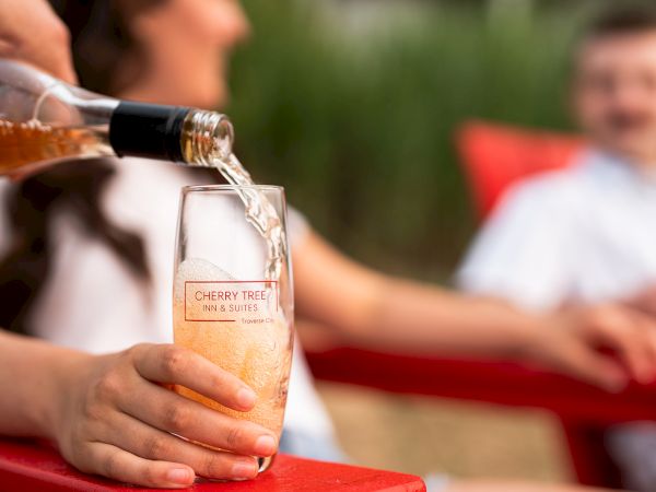 Pouring a glass of pinkish beverage (likely cider or ros&eacute;) into a clear glass, with people in the background at an outdoor setting.