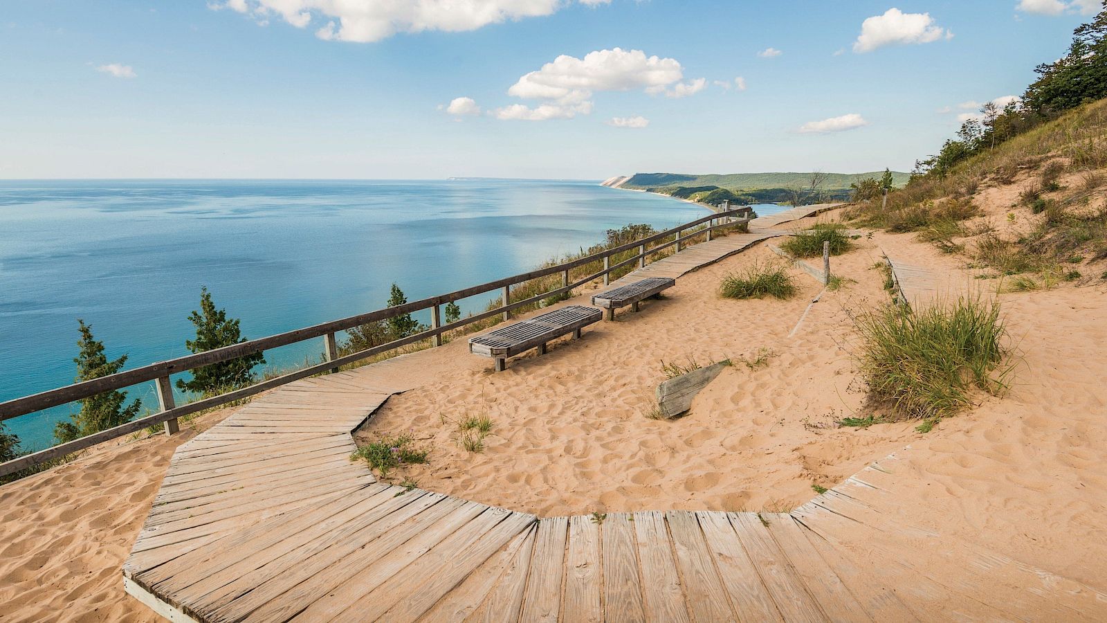 A coastal overlook with a sandy path, weathered wooden boardwalk, and benches facing a calm blue sea under a sunny sky.