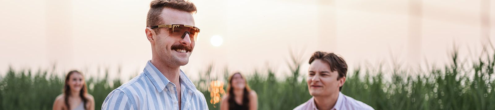 A man in sunglasses holds a beach volleyball while friends jog toward it on a sandy shoreline at sunset.