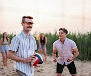 A man in sunglasses holds a beach volleyball while friends jog toward it on a sandy shoreline at sunset.