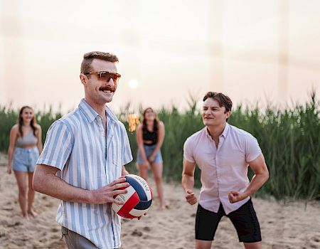 A man in sunglasses holds a beach volleyball while friends jog toward it on a sandy shoreline at sunset.