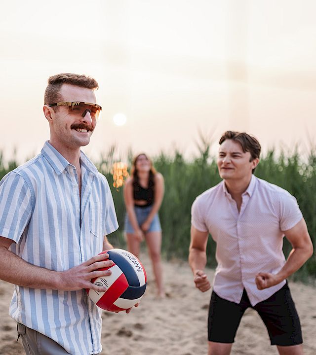 A man in sunglasses holds a beach volleyball while friends jog toward it on a sandy shoreline at sunset.
