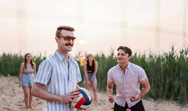 A man in sunglasses holds a beach volleyball while friends jog toward it on a sandy shoreline at sunset.