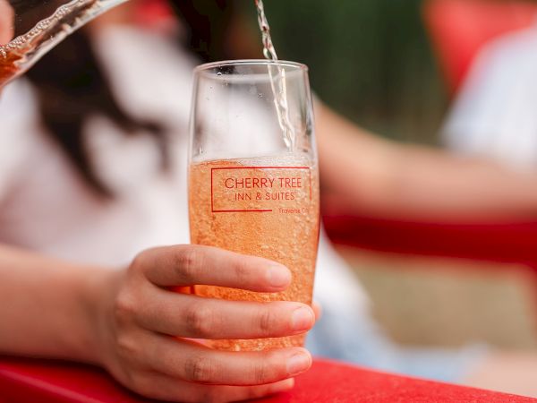 A hand pours into a branded glass labeled &ldquo;Cherry Tree Inn & Suites,&rdquo; with bubbles and a festive red-and-white background.