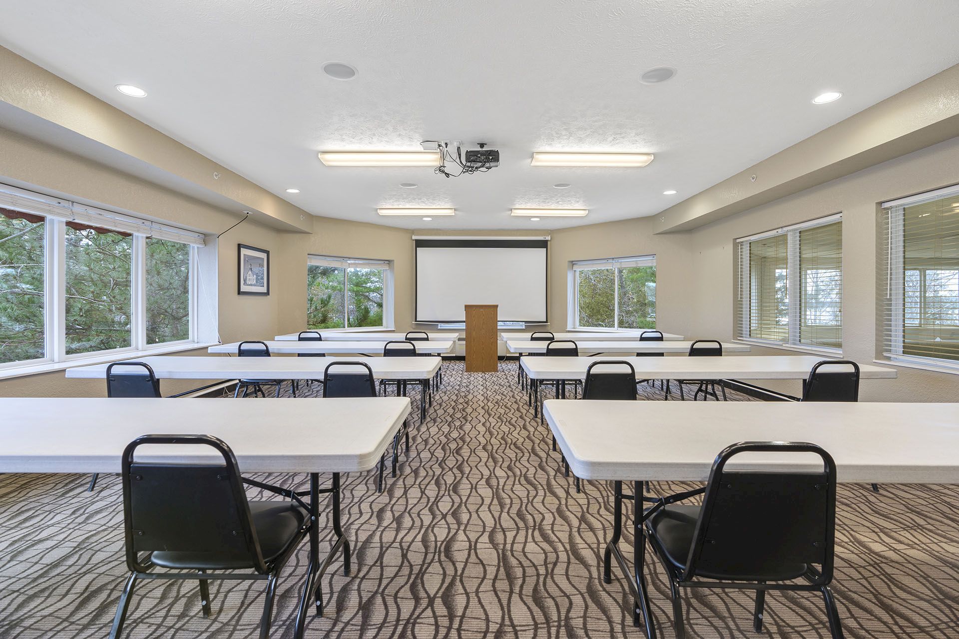 Bright meeting room with long tables, black chairs, a projector screen, and large windows letting in daylight.