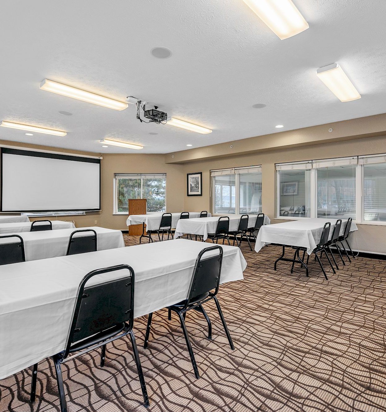 A bright conference room with banquet tables, chairs, a projector screen, and large windows with blinds.