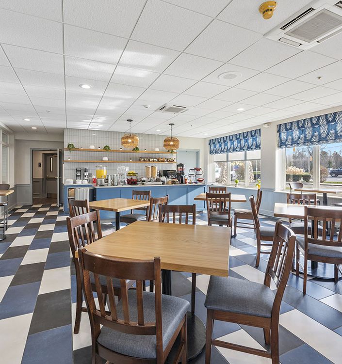 A bright hotel breakfast caf&eacute; with blue-and-white curtains, wooden tables, chairs, and a serving counter near large windows.