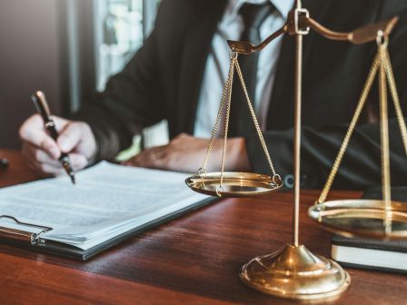 A lawyer writing at a desk beside a gold scale of justice, symbolizing law and balance.