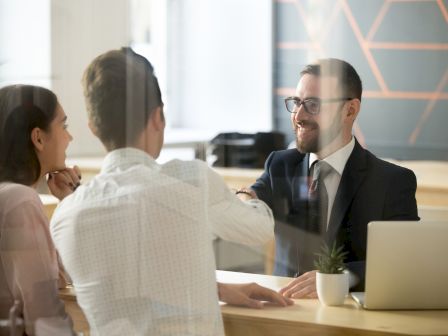 A suited man with glasses chats with colleagues at a conference table during a business meeting in a bright office.