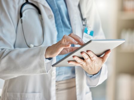 A healthcare professional in scrubs uses a tablet and holds a pen, suggesting they&rsquo;re reviewing patient information.