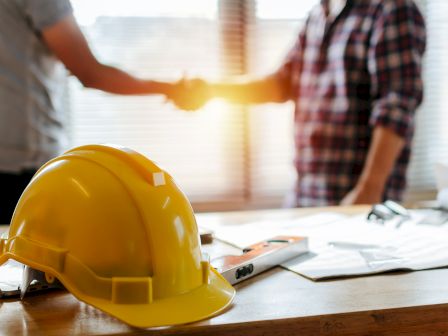 A yellow safety hardhat sits on a desk as two workers in plaid and blue shirts discuss a project plan.