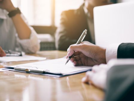 A person&rsquo;s hand writes on paperwork during a business meeting at a table with another person across from them.