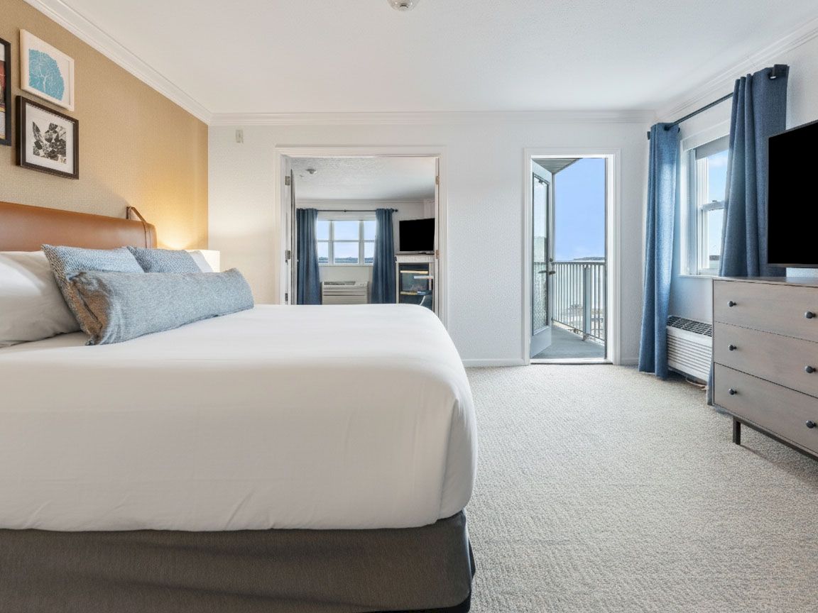 A bright hotel bedroom with a large bed, blue curtains, a dresser and TV, and an open balcony door.