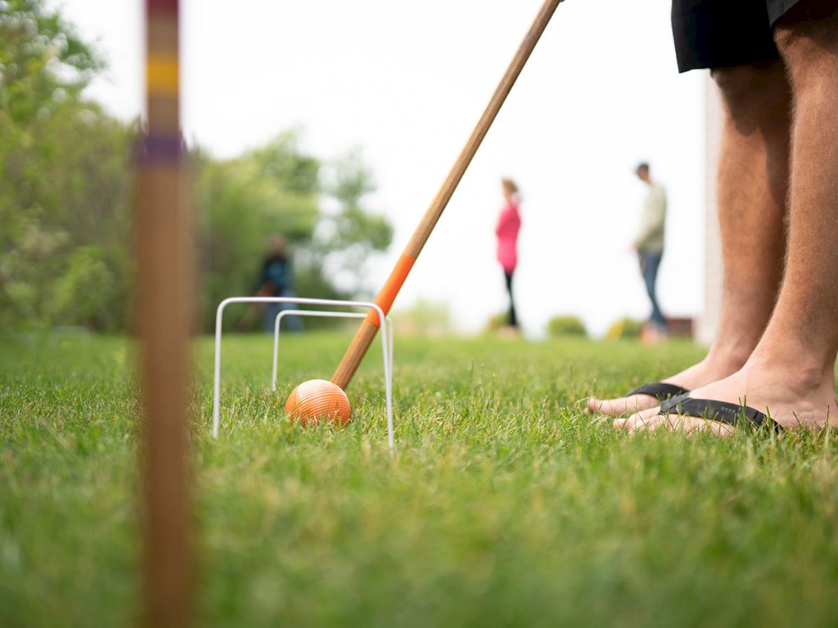 A man plays an outdoor lawn game with an orange ball and mallet, while others stand watching in the grass.