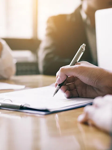 People meet at a table, one hand writing/signing documents with a pen as others look on in soft focus.