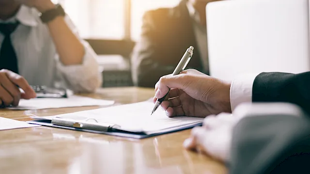 People meet at a table, one hand writing/signing documents with a pen as others look on in soft focus.