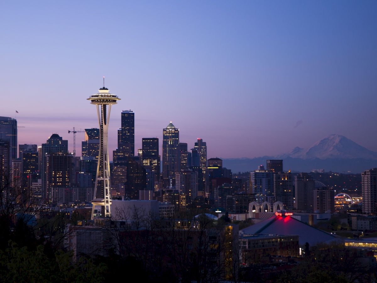 A cityscape at dusk featuring the Space Needle, various skyscrapers, and a distant mountain under a clear, fading sky.