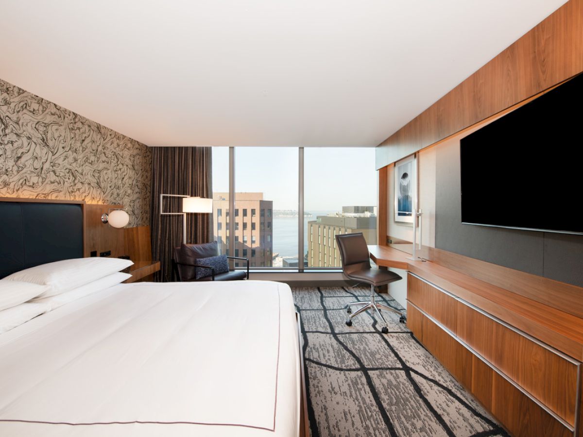 A modern hotel room with a large bed, a wall-mounted TV, a desk with a chair, and a view of city buildings through a large window ending the sentence.