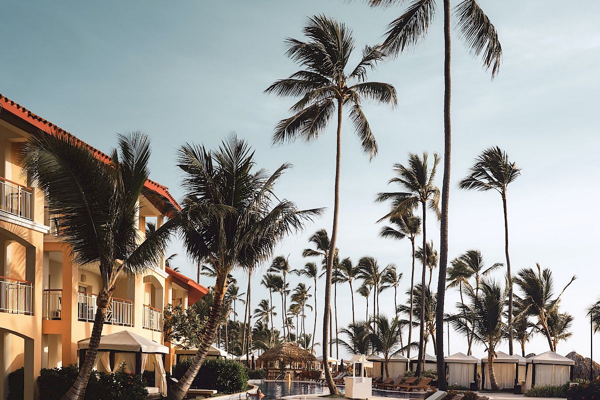 A serene poolside scene featuring tall palm trees, lounge chairs, and a multi-story building with balconies under a clear sky, evoking a tropical vibe.