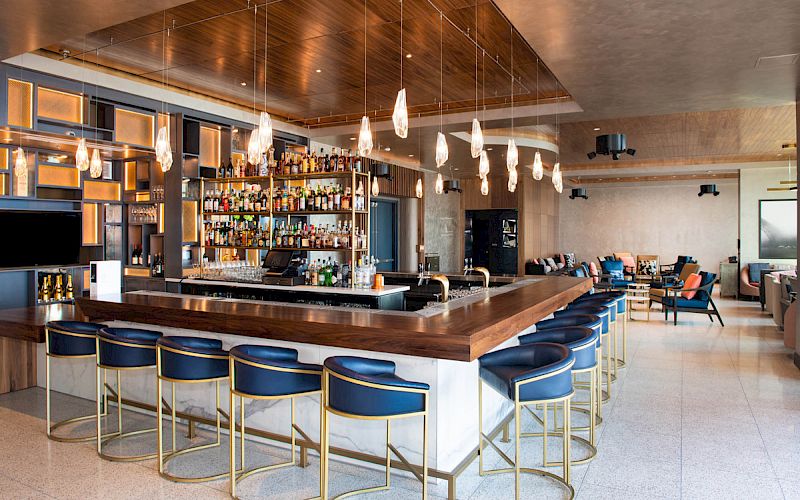 This image shows a modern bar with a sleek design, featuring a long counter, stylish barstools, a well-stocked back bar, and elegant pendant lighting.