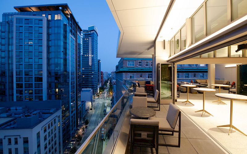 This image shows a modern cityscape at dusk with a view from a balcony, featuring outdoor seating and buildings with illuminated windows.